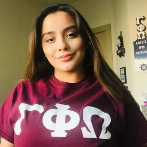 Photo of Maria, wearing a maroon shirt with the Greek Letters Gamma Phi Omega