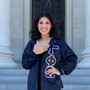 Maya Coy smiling at the administration building with a Gamma Phi Omega jacket doing the organization hand sign
